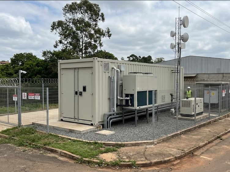 Data center Edge em contêiner bege com unidades de refrigeração, cercado por alambrado com concertina e placas de aviso, ao lado de uma torre de antenas